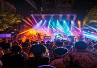 A vibrant concert scene at an outdoor music festival in Barcelona at night, with colorful stage lights illuminating a diverse crowd of enthusiastic fans dancing and cheering. The stage features indie