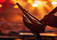 A close-up of a musician's hands playing a Venezuelan cuatro, with the wooden instrument in sharp focus against a blurred background of a concert stage with warm stage lights.