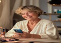 A 61-year-old woman with short hair, sitting at a vanity table with makeup products, smiling naturally at a smartphone camera, soft natural lighting, photorealistic style, no text or logos.