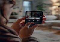 A photorealistic image of a woman's hands holding a smartphone discreetly recording a tense domestic scene, with a blurred background showing a living room. The focus is on the phone screen displaying