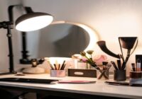 A well-lit makeup tutorial setup with a ring light, mirror, and various cosmetics arranged neatly on a table. The background is a clean white wall. The scene is bright and inviting, with a smartphone