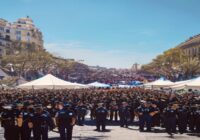 A realistic photo of a large crowd gathered in a public square in Madrid, with police officers in uniform standing in formation and medical tents visible in the background, under a clear blue sky. The