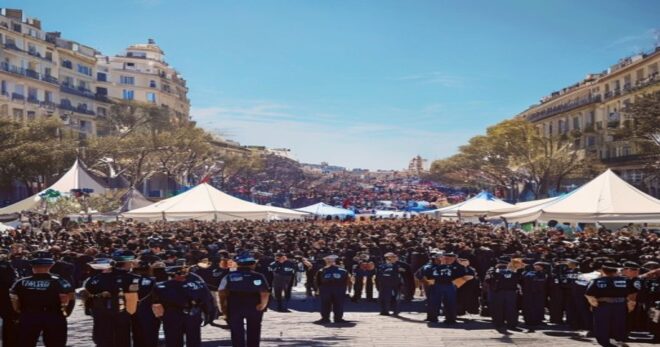 A realistic photo of a large crowd gathered in a public square in Madrid, with police officers in uniform standing in formation and medical tents visible in the background, under a clear blue sky. The