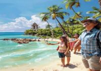 A photorealistic image of a happy tour guide leading a small group of tourists along a sunny beach in Zihuatanejo, with palm trees and turquoise water in the background. The guide is pointing towards