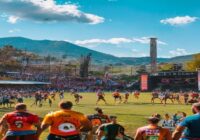 A vibrant scene at a rugby field in Monzón, Spain, during the Fat Rugby festival. Players in colorful jerseys are mid-match, while a stage with musical instruments and speakers is set up nearby. Crow