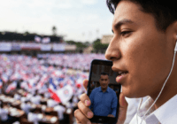 Photorealistic image of a person listening to a political speech on a smartphone with earbuds, with a blurred background of a political rally or parliament. No text or logos.