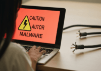 A realistic photo of a person using a laptop with a caution sign about malware, next to a broken electrical cable on the floor, illustrating the analogy between electric shock and dangerous downloads.