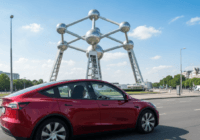 Photorealistic image of a Tesla Model Y driving on a street in Brussels, Belgium, with the Atomium in the background during daytime. The car interior shows a driver with hands loosely on the wheel, an