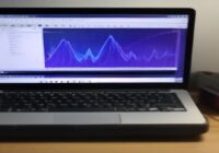 A person's hands typing on a laptop keyboard, the screen displaying the colorful Sonic Pi interface with code and waveform visualizations, in a cozy home studio with headphones and a MIDI controller.